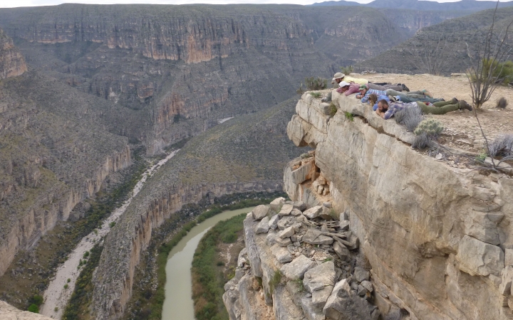 a group of people lay on the ground, peering over the edge of a cliff at the river below